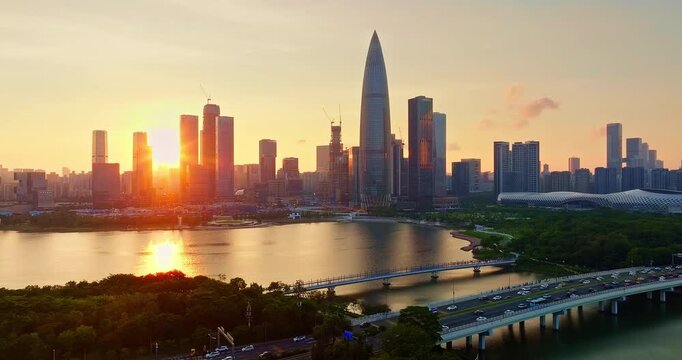 Modern city skyline of Shenzhen, China, featuring high-rise buildings and skyscraper at sunset over a tranquil river with traffic on a bridge and green tree.