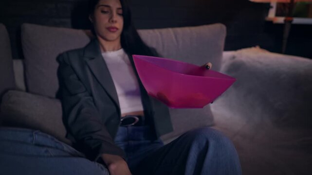 Closeup view of young adult Caucasian woman sitting on the sofa and eating crisps while watching TV. Slow motion of the lady with long dark hair resting at home with bowl of snacks enjoying the film