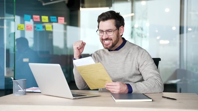 Happy businessman reading letter with great news while sitting at desk at workplace in modern office. Smiling mature professional reacts with pleasant surprise and joy after receiving positive message