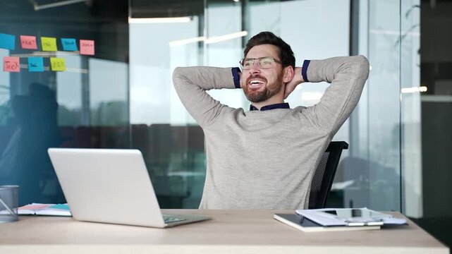 Happy businessman finished work on laptop while sitting at desk in modern office. Smiling satisfied professional leans back in chair puts hands behind head, relaxing after completing task at workplace