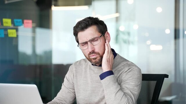 Close up of businessman suffering from ear pain while sitting in modern office. Mature professional touching ear and feeling discomfort, experiencing earache symptoms caused by infection or irritation