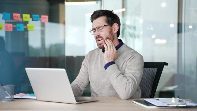 Businessman suffering from toothache while sitting at desk in modern office. Mature professional holding hand on cheek, feeling sharp dental pain and discomfort, reacting to aching tooth during work.