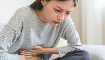 Obraz premium Young woman sitting on sofa and holding her stomach in pain ソファに座り腹痛で苦しそうにお腹を押さえる若い女性
