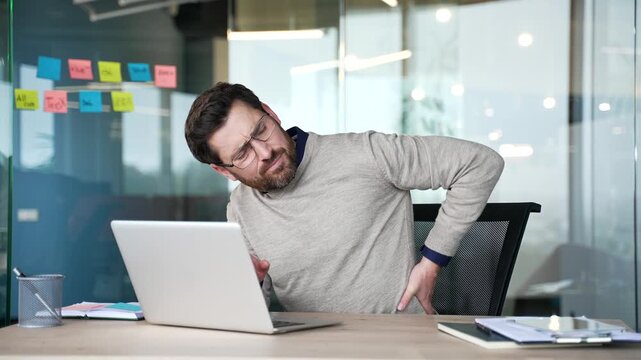 Businessman suffering from back pain while sitting at desk in modern office. Exhausted mature professional massages lower back muscles, feeling tension, fatigue and discomfort caused by prolonged work