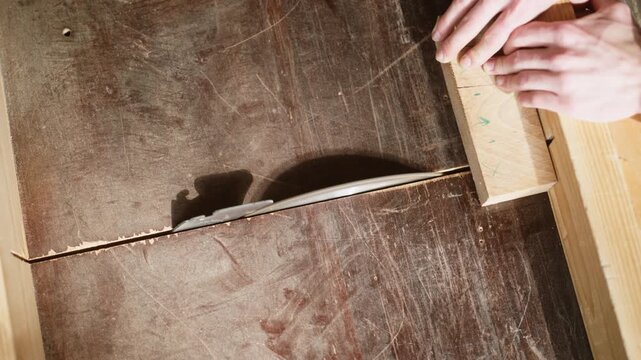 A person's hands guide a wooden block into a circular saw on a table saw. The sharp blade cuts through the wood, creating sawdust.