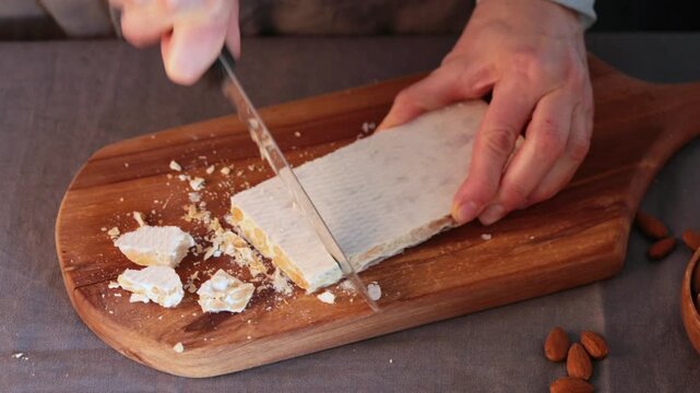 A close-up video of hands using a sharp knife to slice a block of hard white Spanish almond turron on a rustic wooden board with scattered almonds nearby