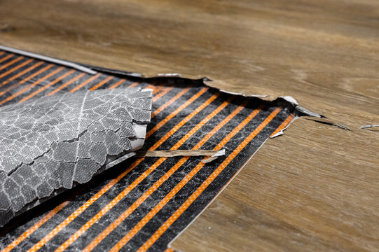 closeup torn flooring revealing foil heating mat. cracked foil patch and exposed thermal element. textured laminate surface with broken backing. diagnostic inspection