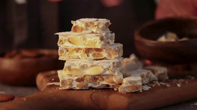 A person's hands carefully put pieces of hard white almond turron into a wooden bowl next to bowl of whole almonds, cutting board and a kitchen knife on a grey tablecloth