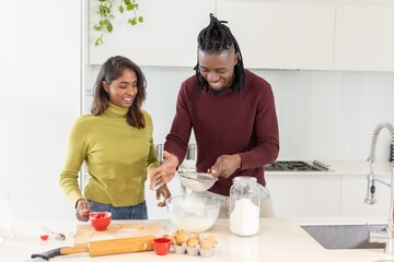 Diverse couple mixing batter at kitchen island using glass bowl, metal sifter, red cups