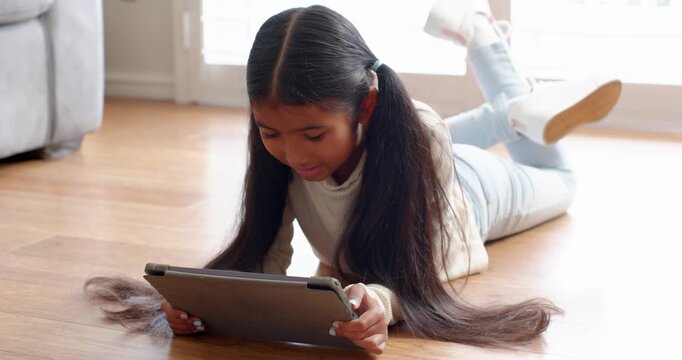 School-age girl propping on elbows and tapping tablet screen for gaming on floor in living room
