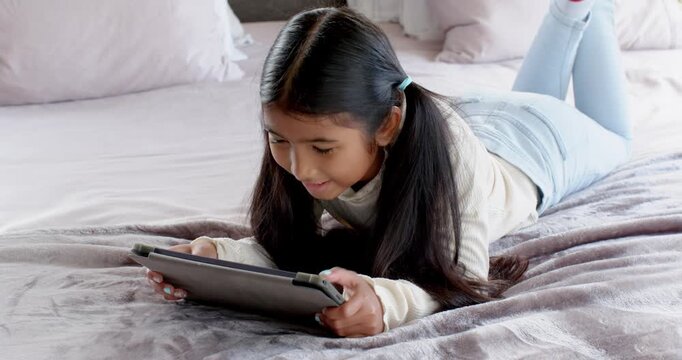 School-aged girl tapping tablet on bed at home pausing to giggle and adjusting angle for learning