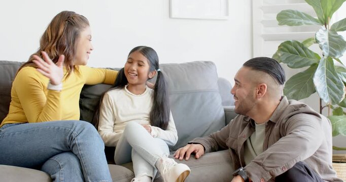 Family sitting on grey sofa in living room, father speaking, mother gesturing and daughter giggling