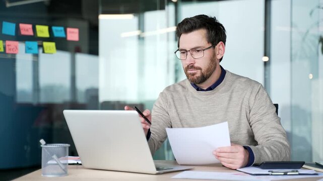 Happy businessman reviewing financial documents and looking at laptop screen in office. Smiling entrepreneur satisfied with results, feeling joy and accomplishment from successful business performance