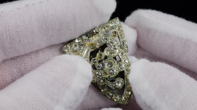 Detailed inspection of jewelry by hands in white cotton gloves. Hands holding vintage art deco dress clip with clear crystals. Ornate silver tone brooch with shimmering baguette and round rhinestones
