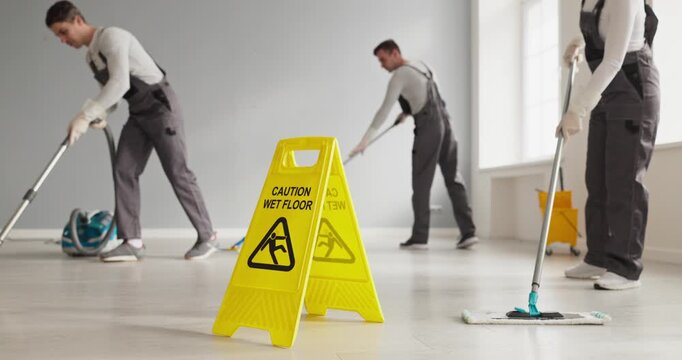 Cleaners janitors team mopping vacuuming warning sign. Yellow wet floor sign in focus as staff sanitize an interior using mop and vacuum to maintain hygiene. Safety at work, hygiene concept.