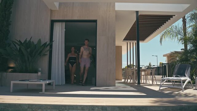 Young couple in swimsuits walking out of luxurious modern villa to poolside. Summer vacation at exclusive resort, sunny day, relaxation, premium lifestyle, travel and leisure concept.
