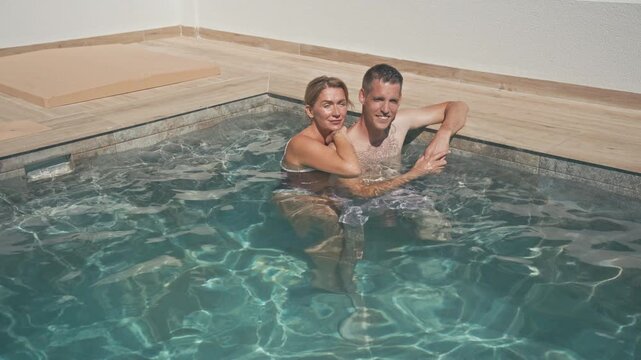 Romantic happy couple relaxing in private swimming pool, enjoy luxurious honeymoon vacation, swimming and embracing during sunny summer day at resort