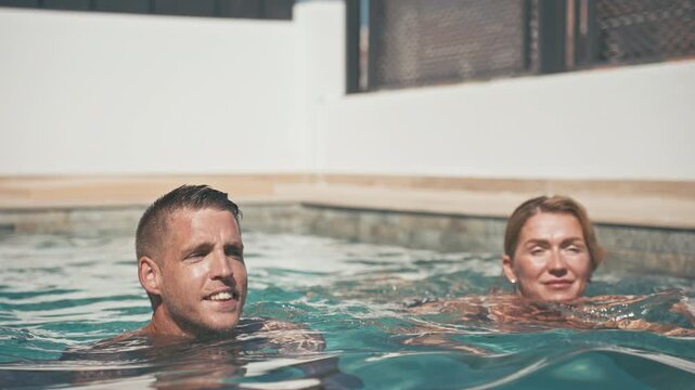 Happy couple enjoying refreshing swim in luxury resort swimming pool. Young man and mature woman relaxing in warm sunlight, sharing joyful moment, summer vacation, leisure and lifestyle concept.