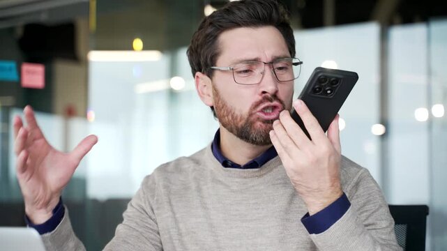 Close up of frustrated businessman arguing on smartphone while sitting at desk in modern office. Angry mature professional reacting emotionally during heated phone conversation and showing irritation