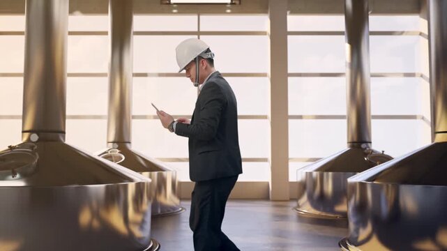 Side View of Asian Businessman Use Tablet While Walking at Modern Stainless Steel Brewing Vats in Large Scale Beer Manufacturing Plant