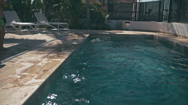 Young boy cannonballing into swimming pool during hot summer day, splashing turquoise water around poolside in tropical backyard with lounge chairs