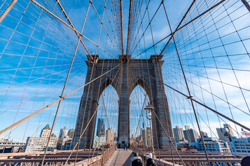 Fototapeta premium Symmetrical view of the Brooklyn Bridge pedestrian walkway leading toward the stone tower, framed by suspension cables and city buildings under a clear blue sky in New York City, USA. 