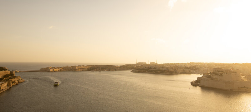 The Grand Harbour glows under a warm sunset, with Fort St. Angelo standing as a sentinel over the tranquil waters as a ferry crosses the historic port.