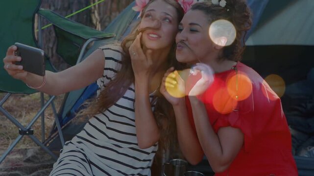 Two women taking social selfie, left woman holding phone, posing faux mustaches, bokeh floating