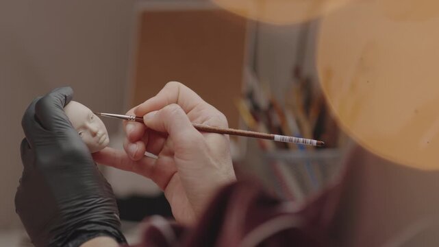 Closeup of skilled artist painting facial details on handcrafted doll head using fine brush, showing precision, creativity and attention to detail in workshop