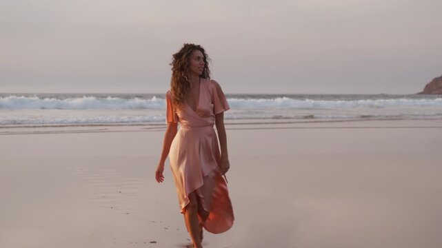 Zoom in closeup shot of joyful woman in pink dress walking on sandy beach and looking at camera during golden sunset