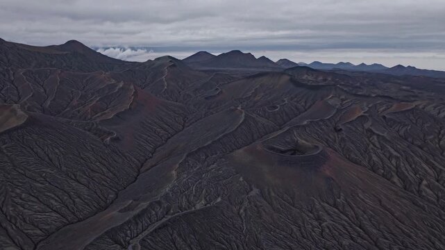 Aerial volcanic desert under overcast sky, remote badlands with braided lava channels, steep ashscoured ridges, scattered cinder cones, muted cool palette, distant mountain silhouettes, cinematic