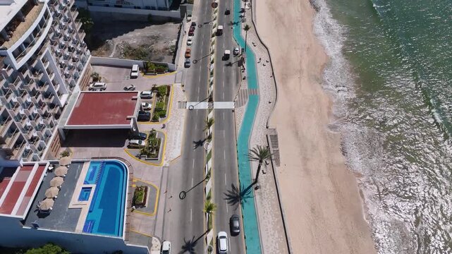 Vista a&eacute;rea con dron del malec&oacute;n de Mazatl&aacute;n, Sinaloa, M&eacute;xico, mostrando una avenida costera con tr&aacute;fico vehicular moderado junto a la playa. Se observan hoteles frente al mar con albercas.