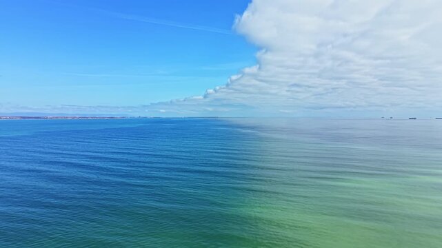 A wide view shows the ocean extending to the horizon under a bright sky. Waves move gently, and there is a noticeable change in water color along the surface near the shore.