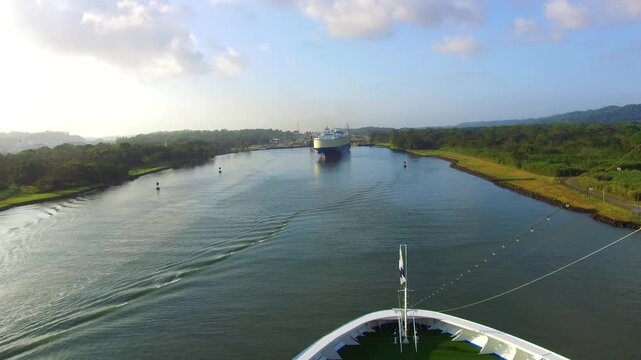 A large cargo ship enters and moves through the locks of the Panama Canal. Surrounding buildings and green areas can be seen. Workers manage the process as the vessel passes through.