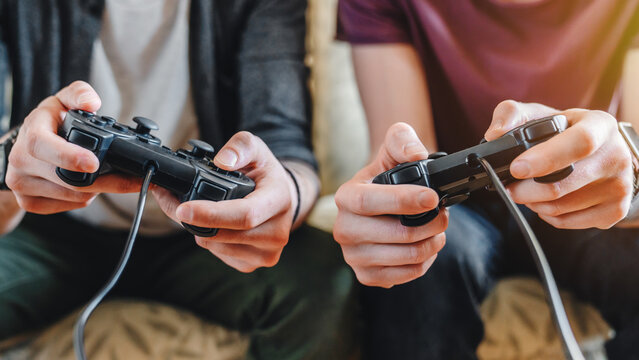 Cropped image of young men playing video games on sofa at home, two friends with controllers having fun, indoor leisure activity, gaming lifestyle, friendship and relaxation concept