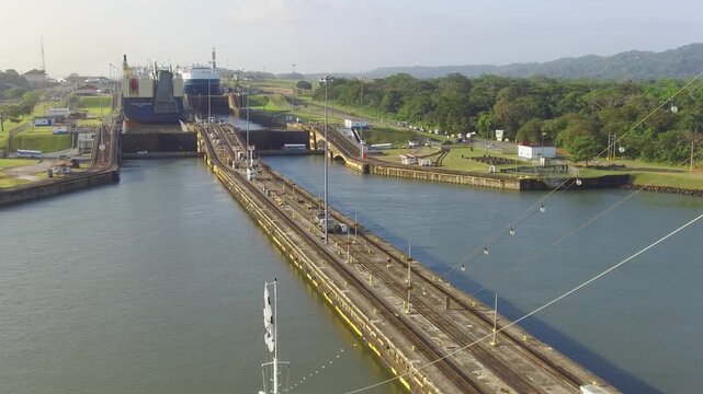 A large cargo ship enters and moves through the locks of the Panama Canal. Surrounding buildings and green areas can be seen. Workers manage the process as the vessel passes through.