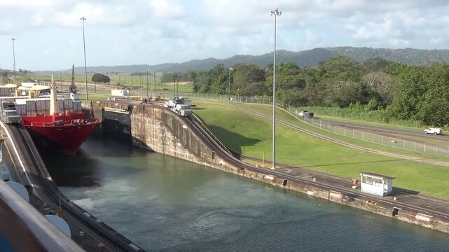 A large cargo ship enters and moves through the locks of the Panama Canal. Surrounding buildings and green areas can be seen. Workers manage the process as the vessel passes through.