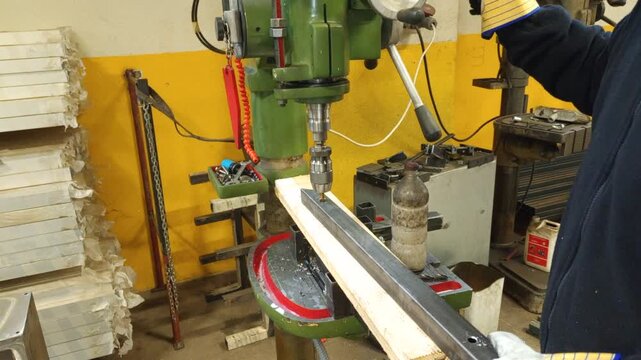 Vertical drilling machine boring a hole in a square metal profile in a workshop.
