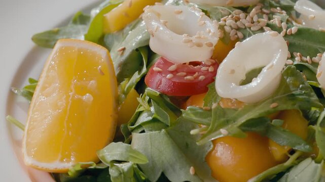 Close-up of fresh salad with mango, arugula, cherry tomatoes, citrus and squid rings rotating on white plate. Elegant restaurant dish presentation with olive oil and sesame dressing. Mediterranean