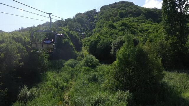 Aerial footage captures a chairlift moving through a lush green forest, eventually revealing a charming mountain resort nestled in a scenic valley on a bright, sunny summer day.


