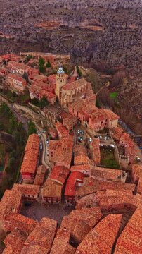 Aerial view shows a village with red rooftops and narrow streets. Stone buildings are scattered among trees. The village is set in a rocky landscape under clear sky.