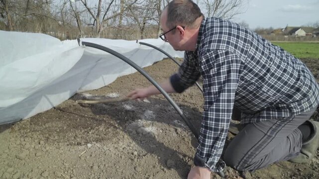 Man in plaid shirt kneels to prepare soil under protective cover, using a wooden tool to cultivate the earth in a garden setting