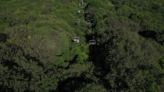 Aerial footage captures a chairlift moving through a lush green forest, eventually revealing a charming mountain resort nestled in a scenic valley on a bright, sunny summer day.

