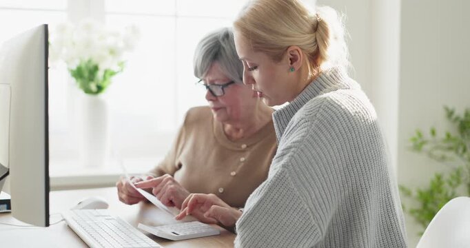 Daughter helps senior mother with bills budgeting. At home they review finances, plan payment schedules on a computer, track expenses and savings, prepare taxes. Family help and finance trust.
