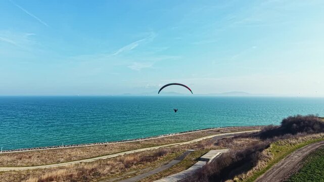 A person is paragliding above the ocean during the day. The sky is clear and the water looks blue. The landscape below has grass and pathways along the coast.