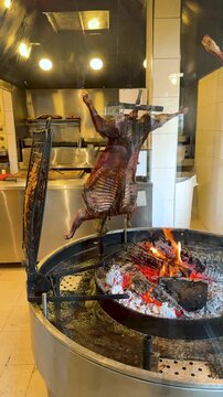 Traditional Patagonian Cordero al Palo, a whole lamb slow-roasted over an open wood fire on a cross-shaped spit in a professional restaurant kitchen. Argentine Asado style whole lamb barbecue