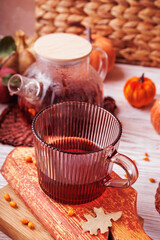 Fototapeta premium Glass cup with red herbal tea and teapot placed on rustic wood with autumn foliage, apple and pumpkins in background