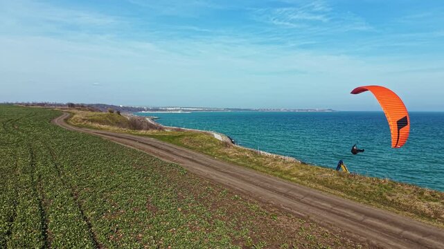 A person is paragliding above the ocean on a clear day. Fields stretch out on one side, and the blue water meets the sky in the distance. The coastline can be seen along the horizon.