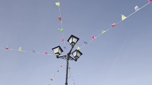 Panning shot of street lamp post with colorful bunting flags fluttering against clear blue sky in town square