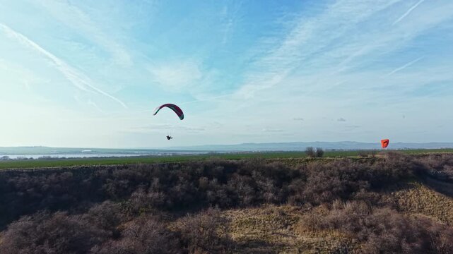 Two paragliders glide through the sky above fields and hills. One is flying high while the other is slightly lower. The scenery includes green land and a distant view of mountains on a clear day.